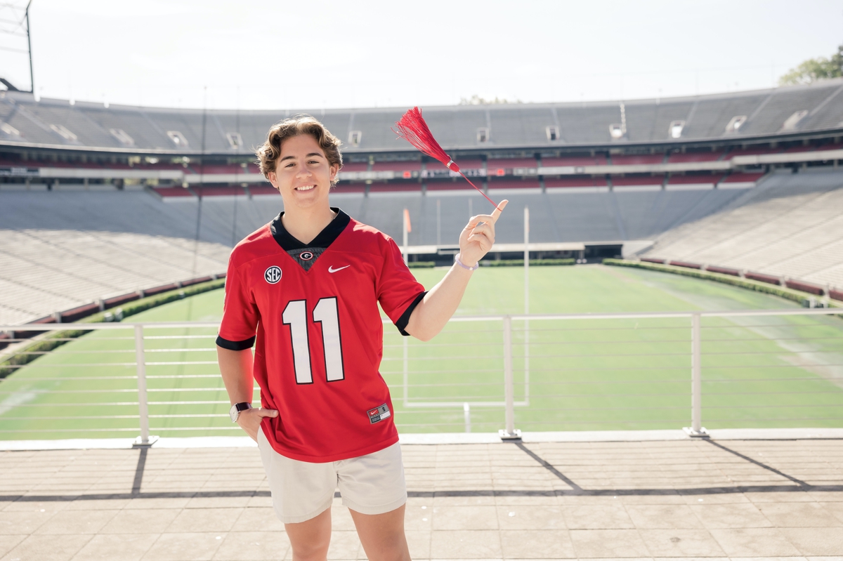 A man wearing a red number 11 football jersey spinning his graduation tassel on his finger in front of the UGA football field