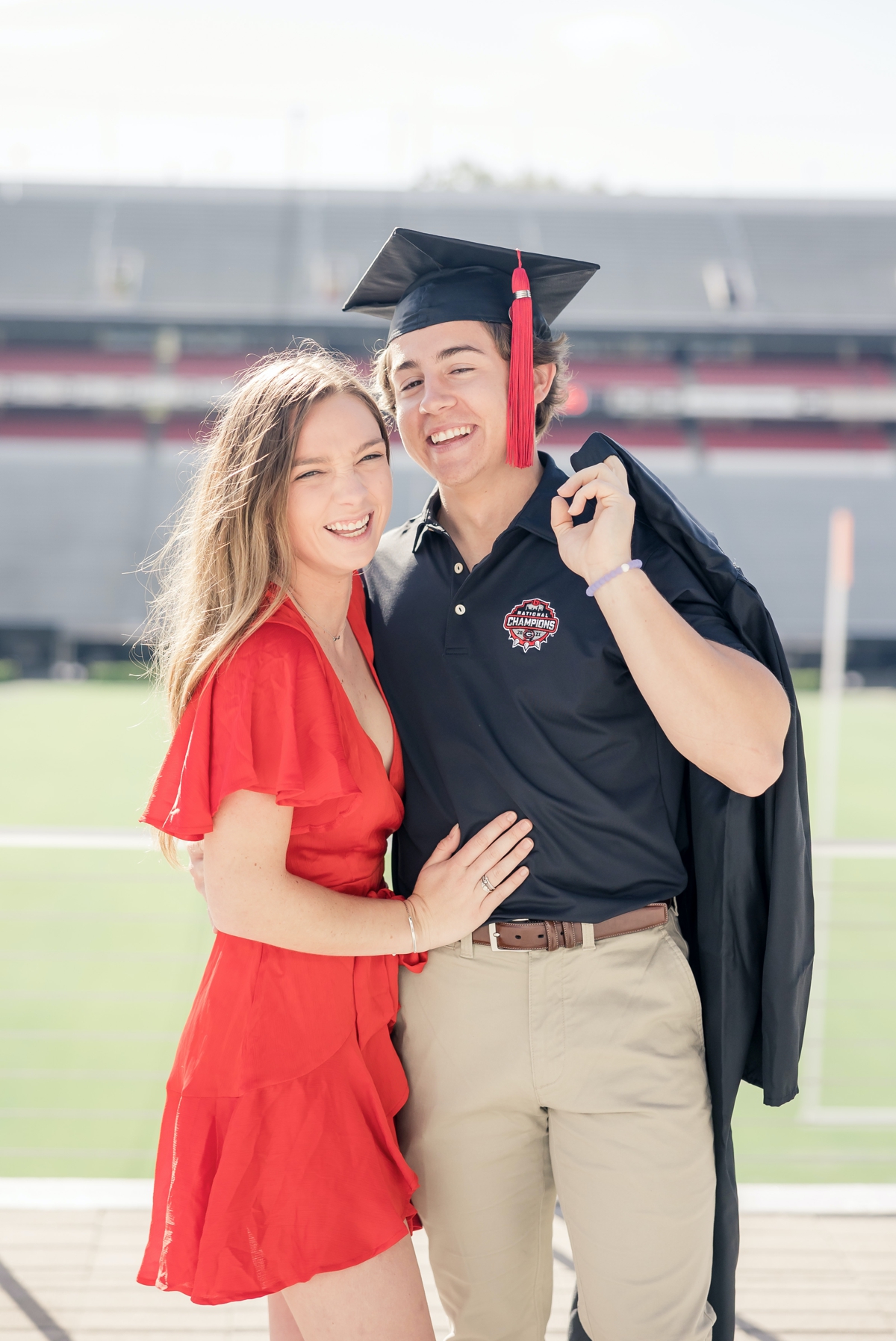 A man and his girlfriend stand on the UGA football field smiling while he wears his graduation cap and has his gown thrown over his shoulder
