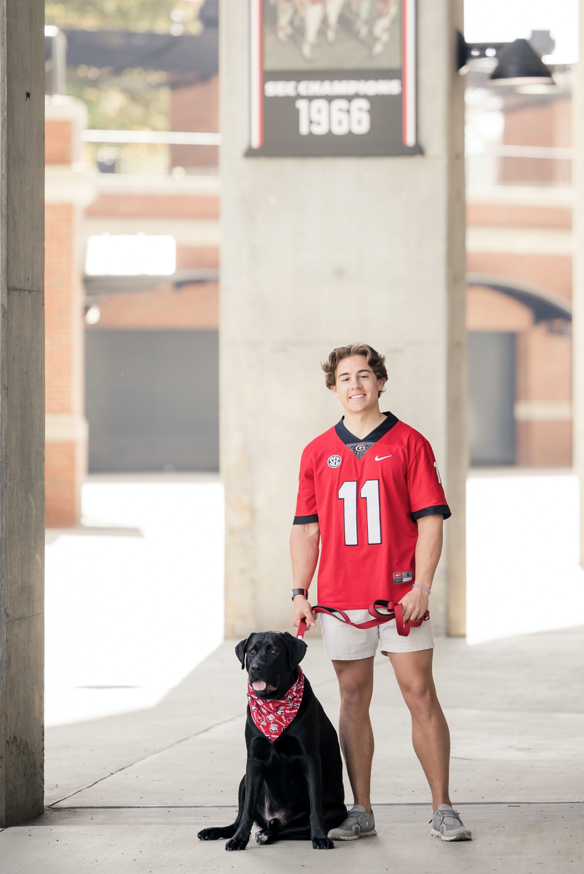 A man in a red jersey stands in the football stadium with his dog during his graduation photo session at UGA