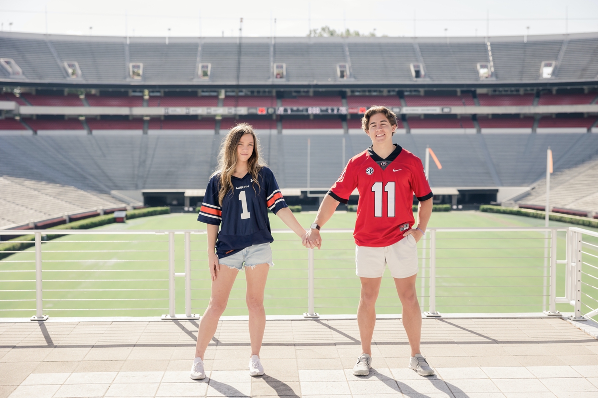 A man and his girlfriend holding hands in front of the UGA football field. He is wearing a red jersey and she is wearing a navy jersey.