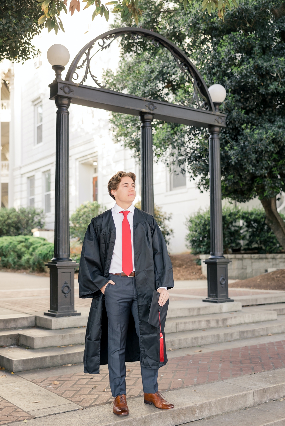 A UGA graduate stands in his gown in front of the campus entrance while looking off into the distance and holding his cap in one hand