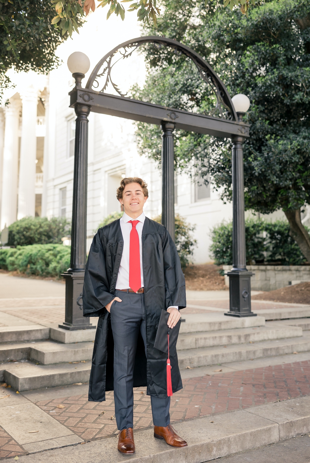 UGa graduate standing smiling in front of the entrance to campus wearing his black graduation robe and holding his cap in one hand during his graduation photo session
