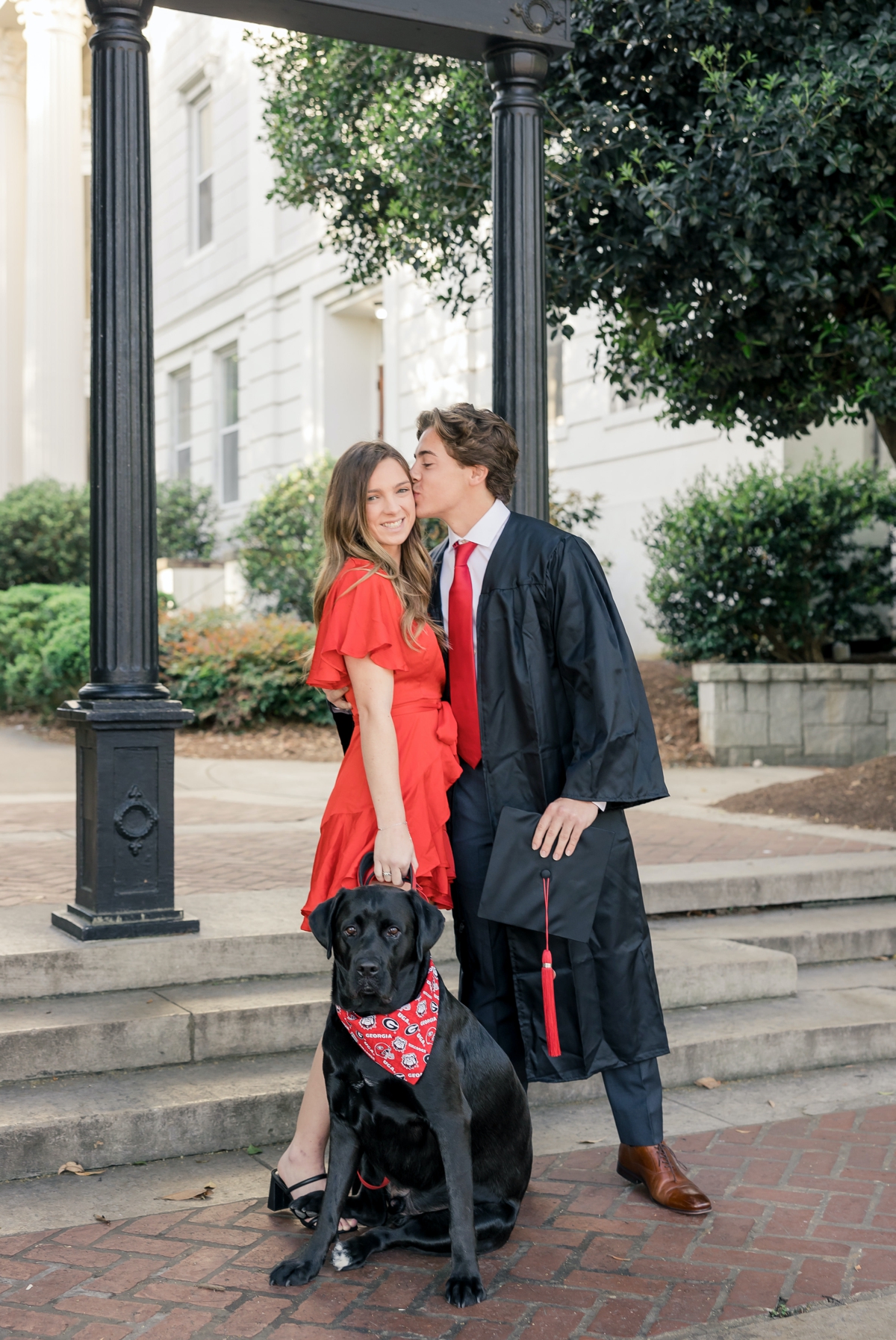 A man kisses his girlfriend on the cheek while dressed in a graduation robe and her holding their dog's leash.