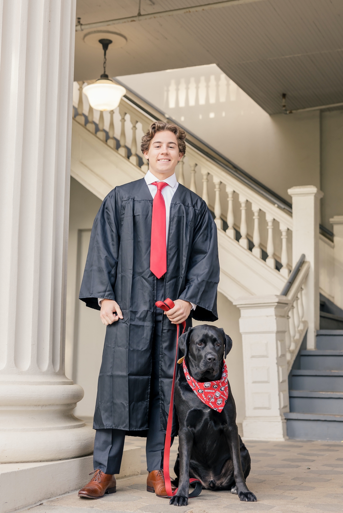 A UGA grad smiling in his graduation gown in front of marble stone steps holding his dog's leash.