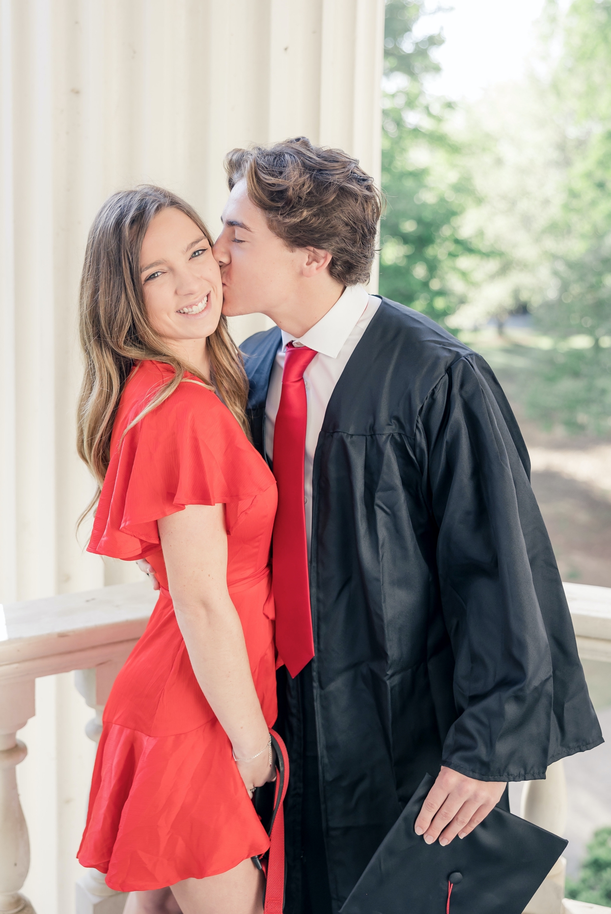 UGA graduate kisses his girlfriend on the cheek while they both stand on the porch of a campus building.