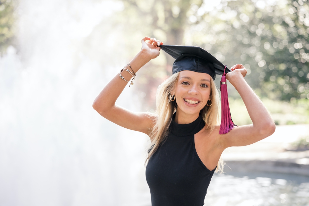 Woman holding her college graduation cap in both hands on her head while a fountain goes off behind her.