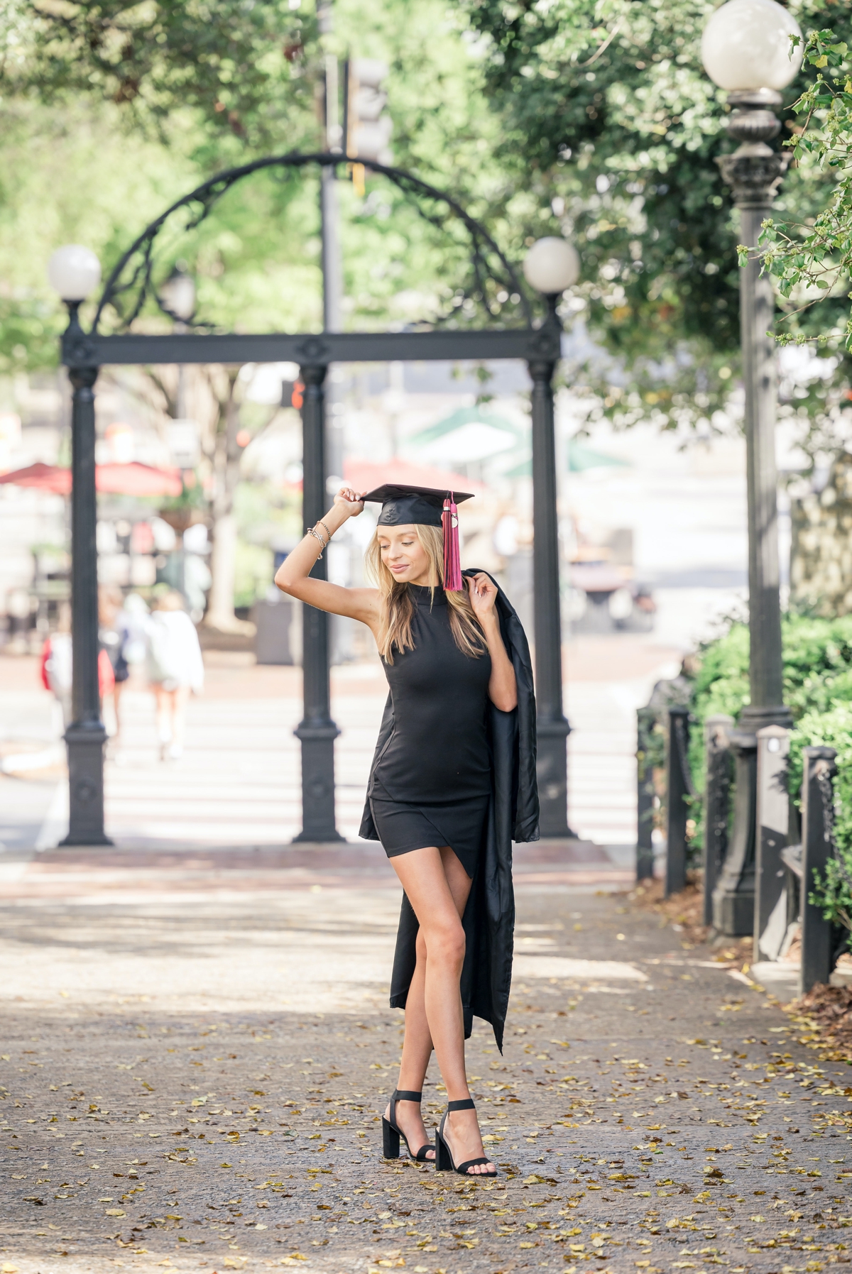 A woman in a black cocktail dress holding her graduation cap on her head while she holds her gown in the other over her shoulder