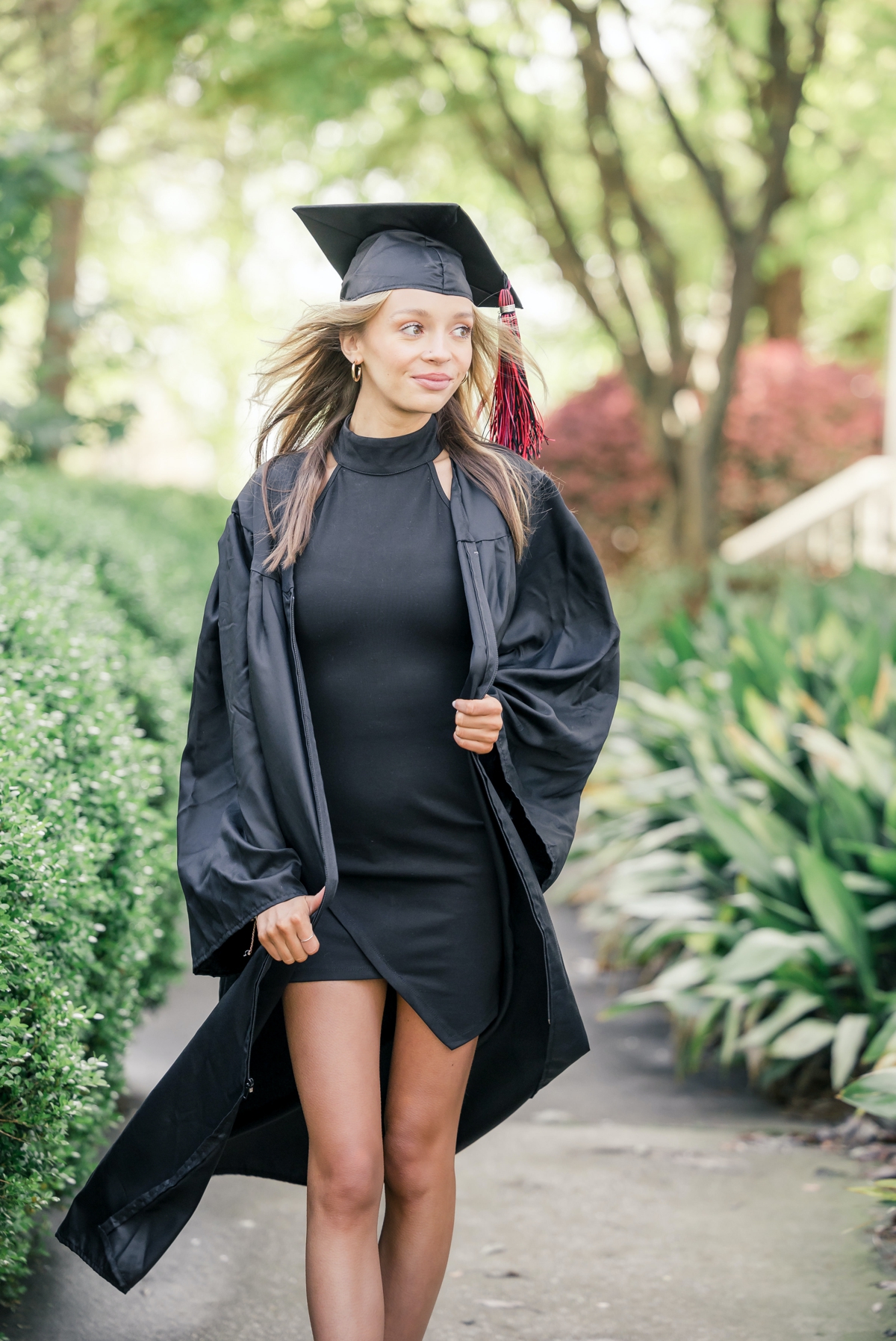 Woman walking through a garden looking off into the distance while she wears her college cap and gown.