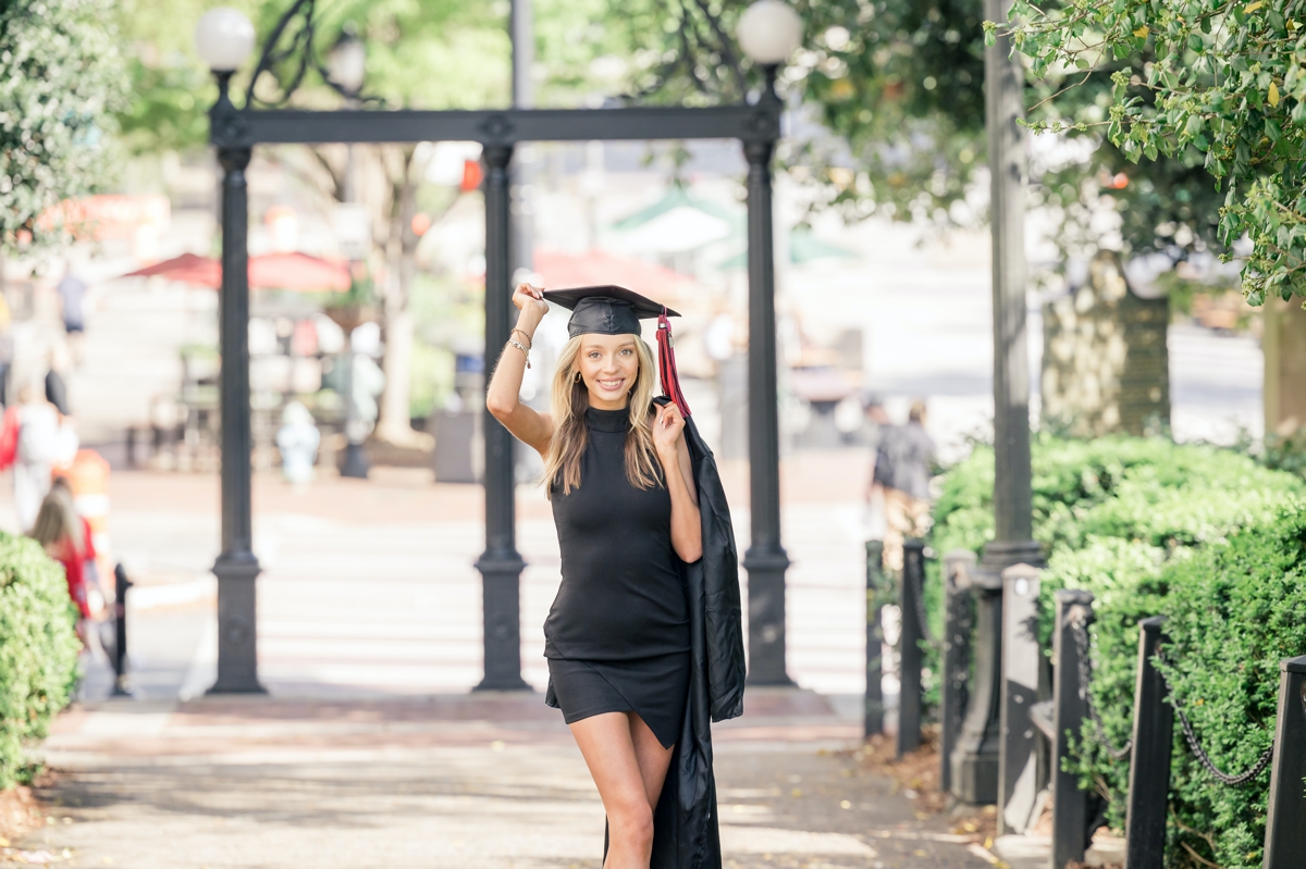 UGA graduate smiling at the campus entrance while she holds her cap on with one and and her gown in the other over her shoulder.