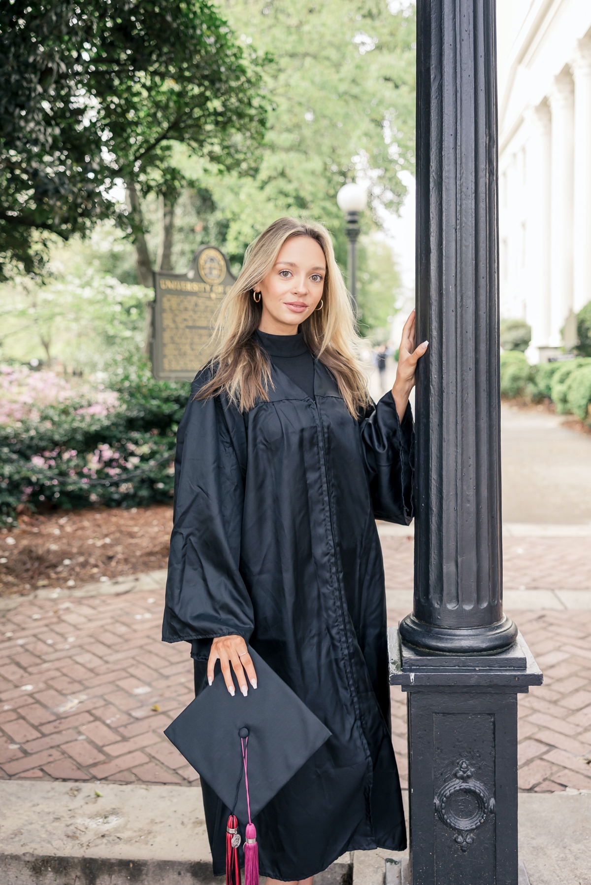 UGA graduate in her black gown holding her graduation cap in one hand and the other hand is placed on a post at the entrance to campus