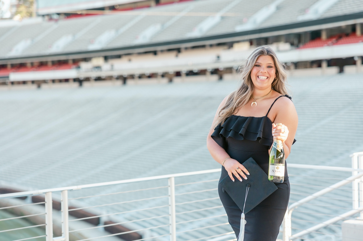 A college senior holding out a bottle of champagne in the football stadium during her senior session.
