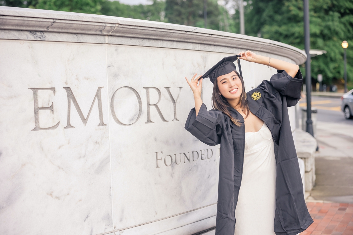 An Emory graduate in front of the campus entrance with both hands on her graduation cap.