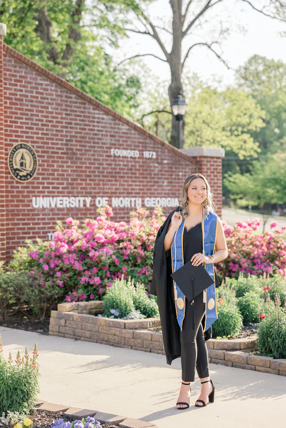 University of North Georgia graduate holding her cap and gown in front of the brick entrance to campus.