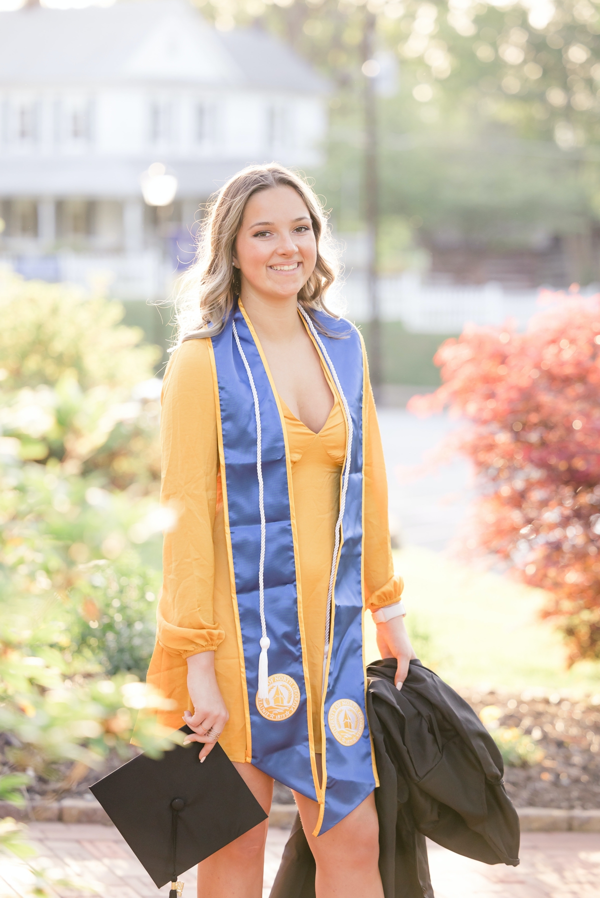 UNG graduate in a yellow dress wearing her graduation stole and cords while holding her cap in a garden on campus.