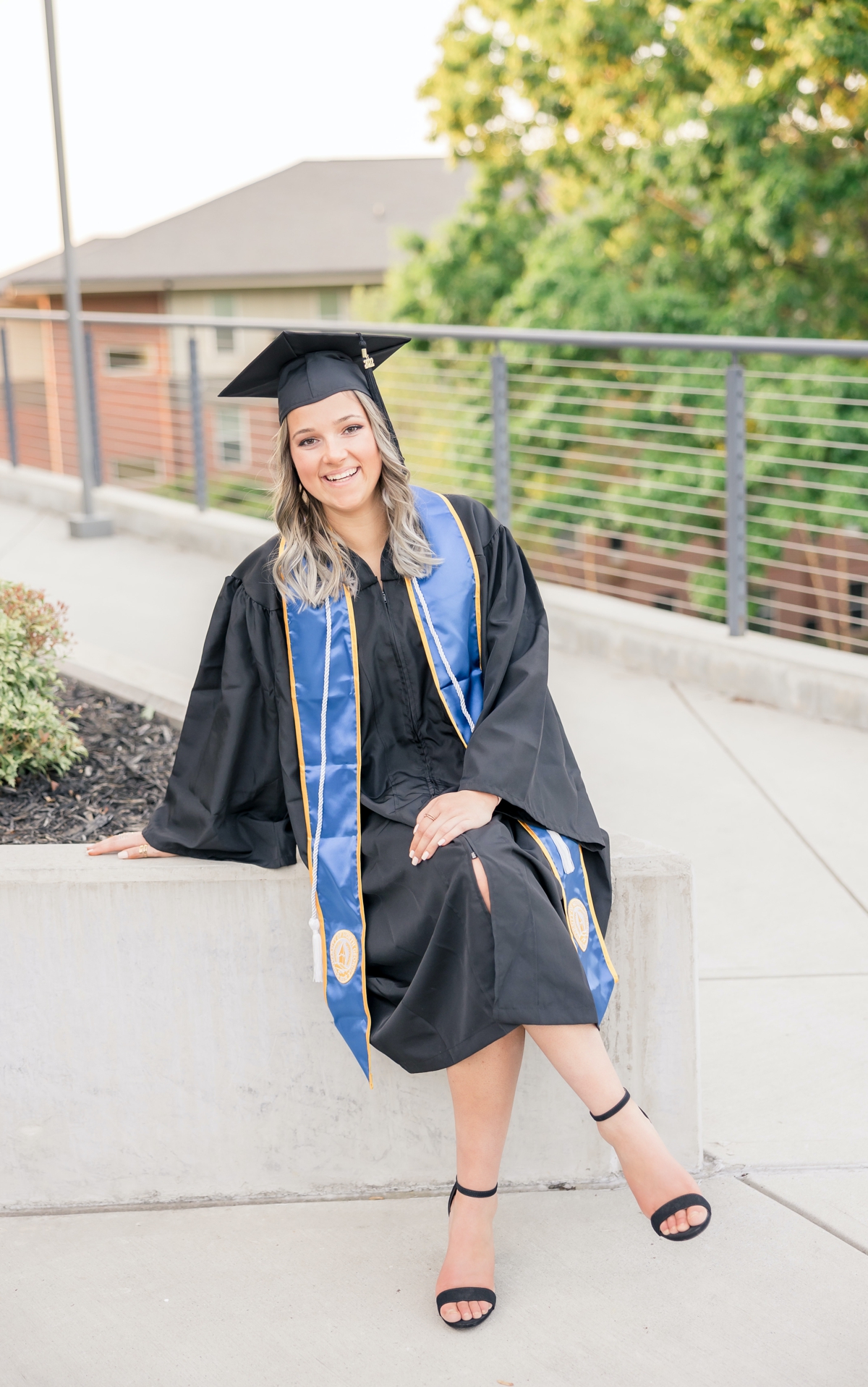 A woman sitting on a concrete ledge in her cap and gown during her graduation session.