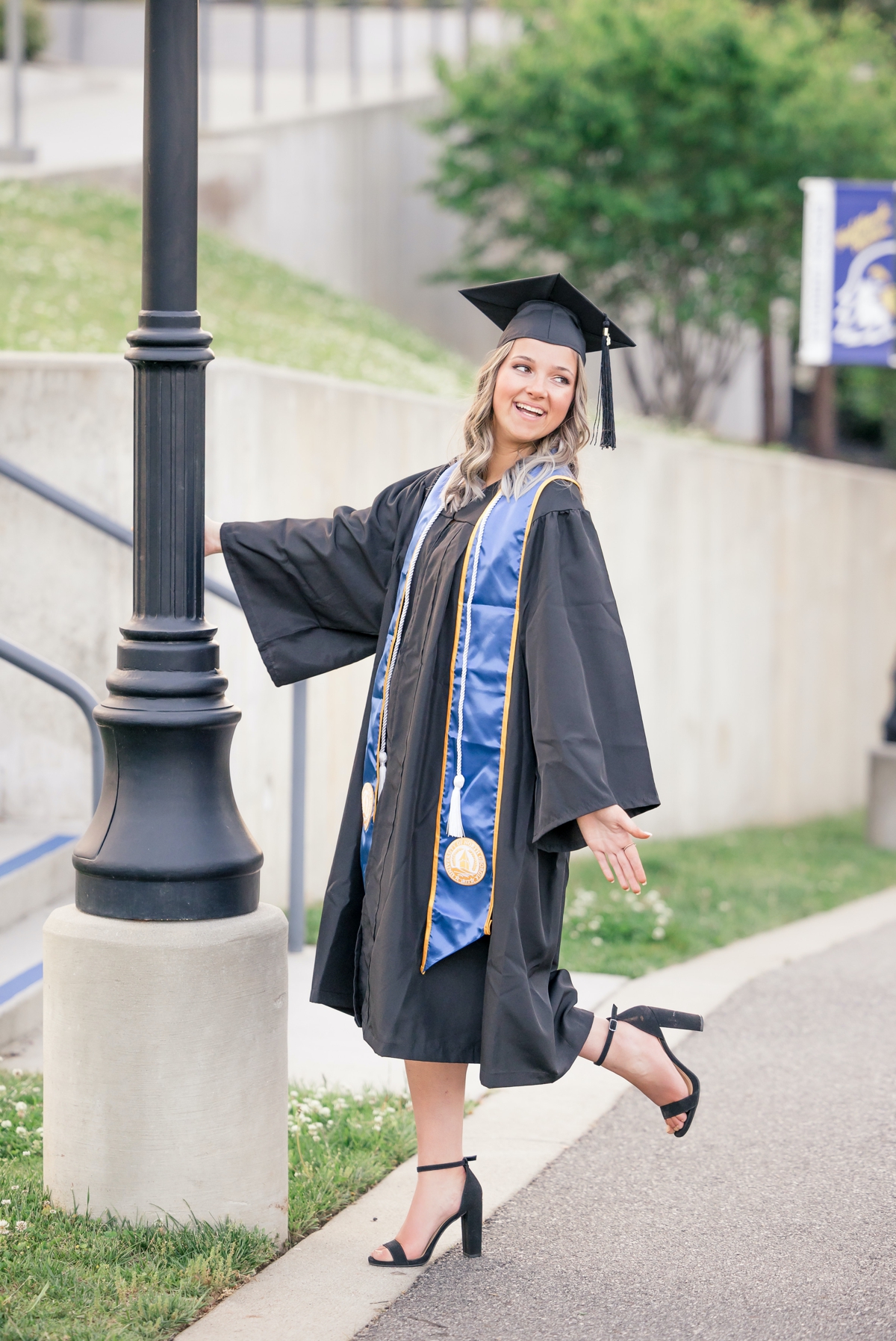 A woman in her University of North Georgia cap and gown laughing over her shoulder while she holds on to a light pole.