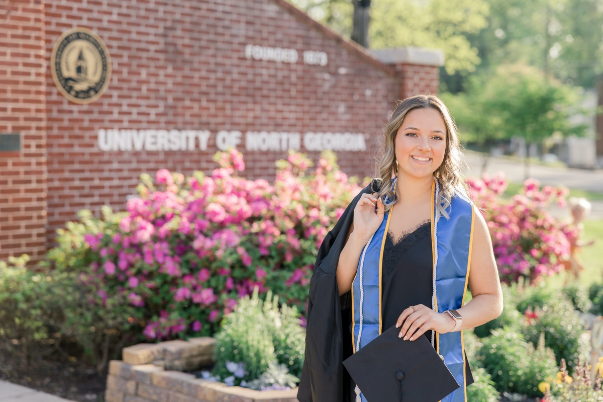 A woman holding her graduation cap and gown at the entrance of the University of North Georgia.