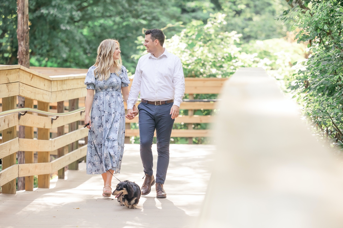 Julia and Taylor walking hand in hand with their dog while they look at each other during their engagement session.