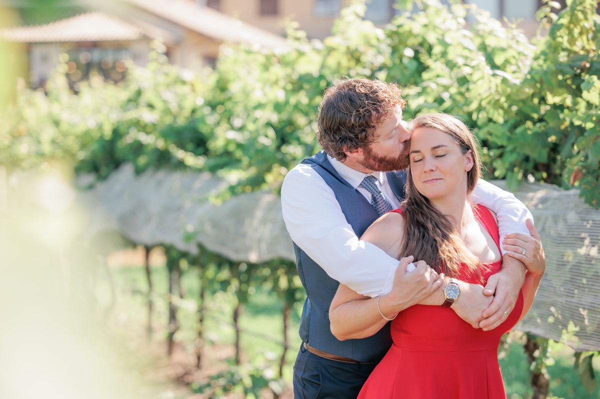 Matthew kissing Rachel on the cheek while he hugs her from behind while standing in the vines during their engagement session.