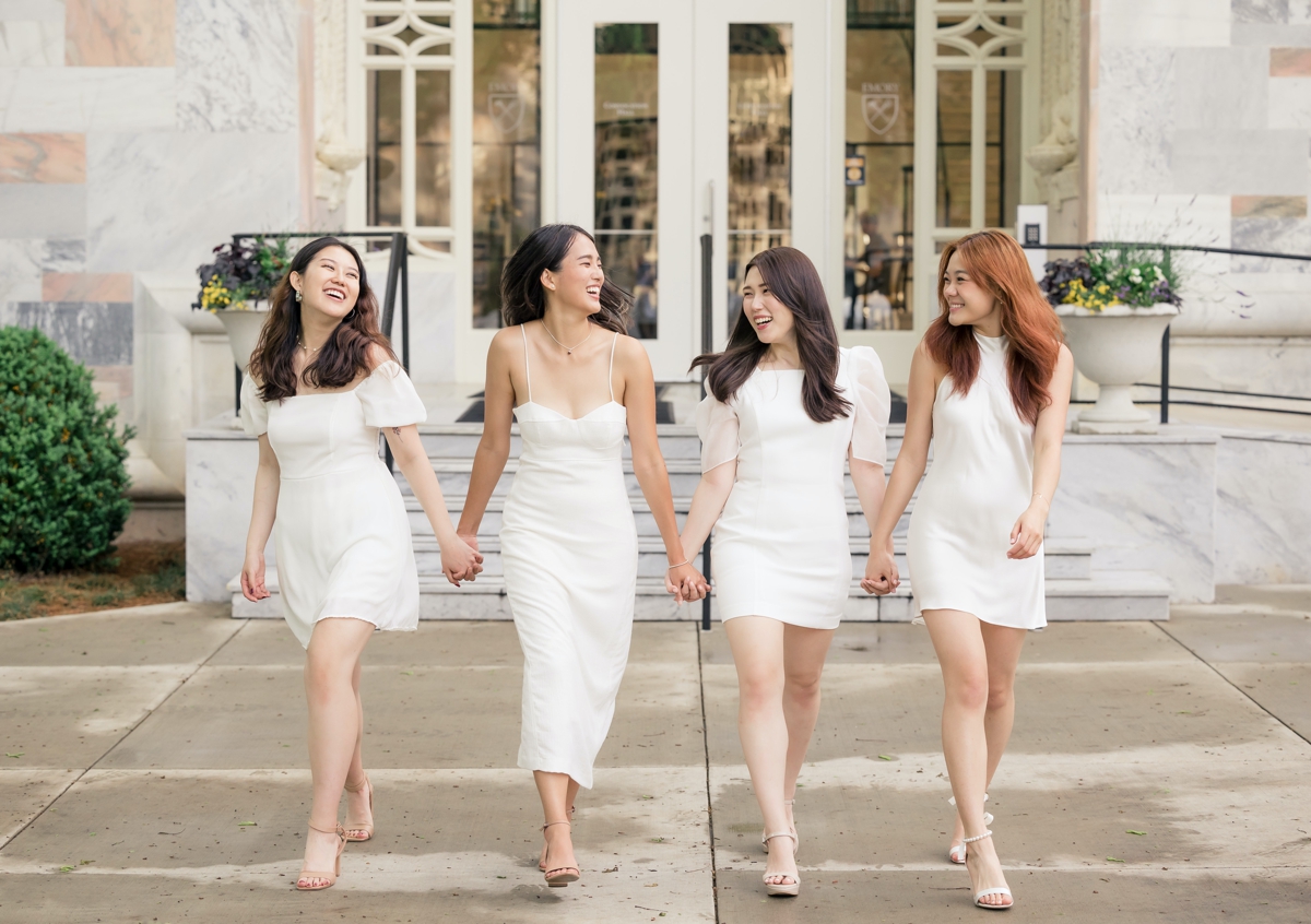 Four women walking hand in hand in white cocktail dresses smiling at each other during their Emory University senior session.