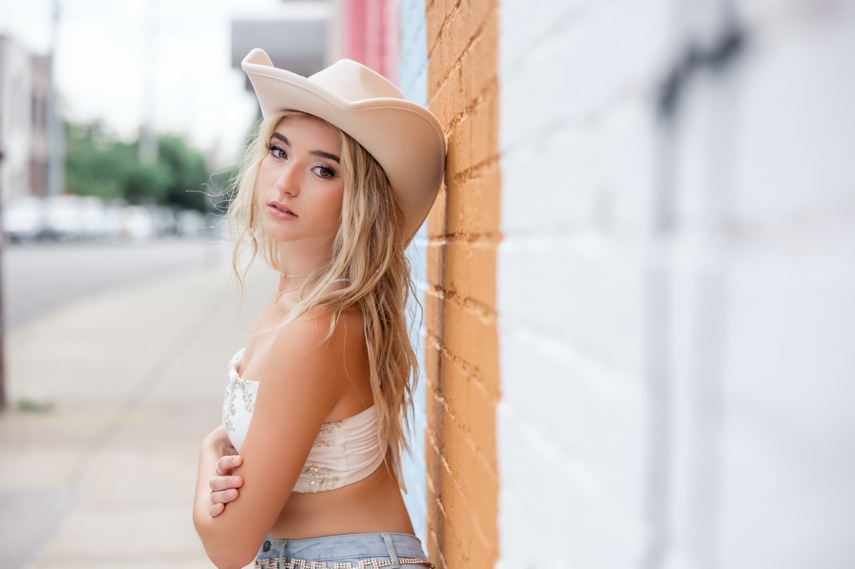 A blonde haired senior girl leaning against a mural wall in a white cowboy hat during her senior session.