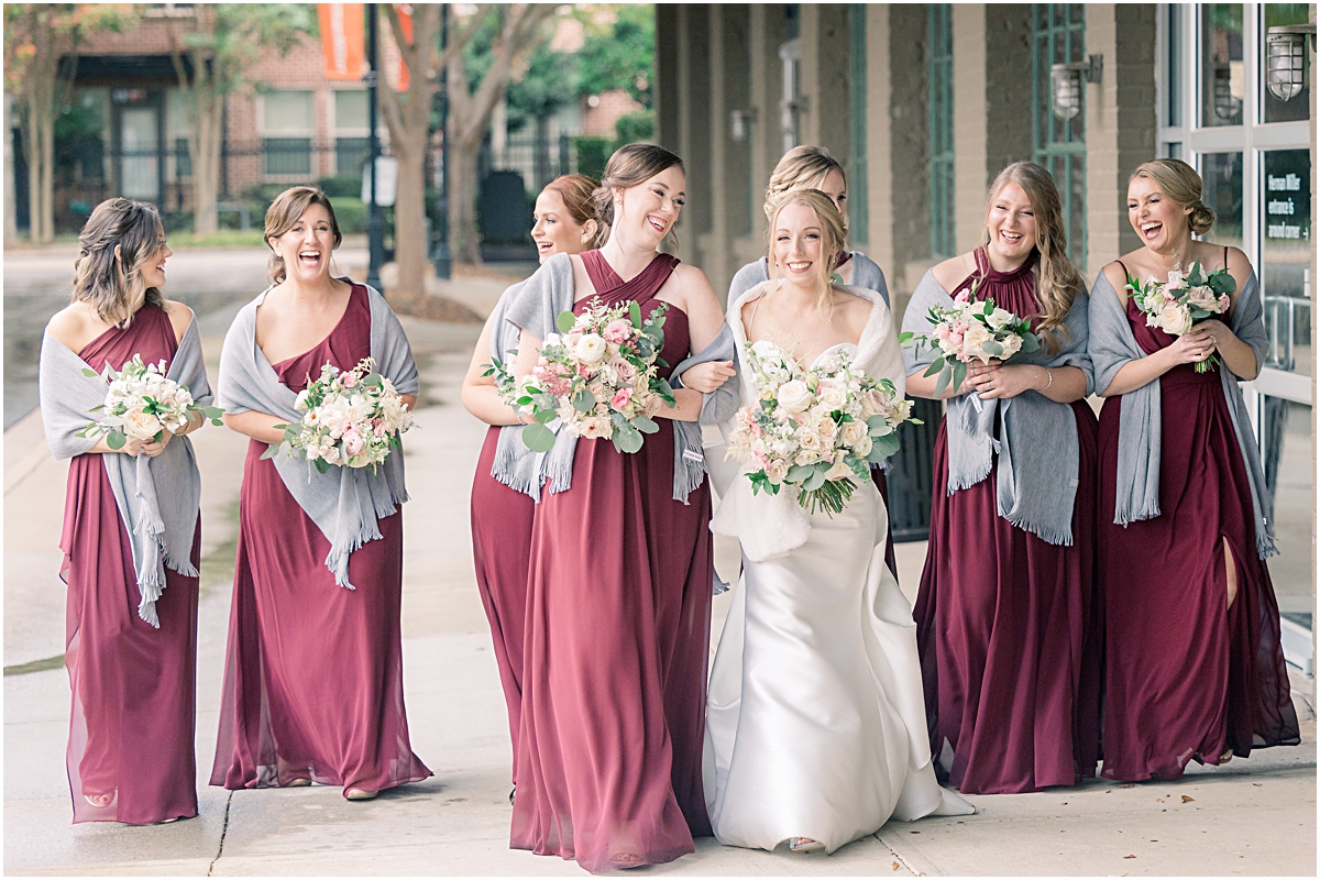bride walking and laughing with her bridesmaids in their maroon bridesmaid dresses with slate blue scarfs around their arms holding their wedding bouquets