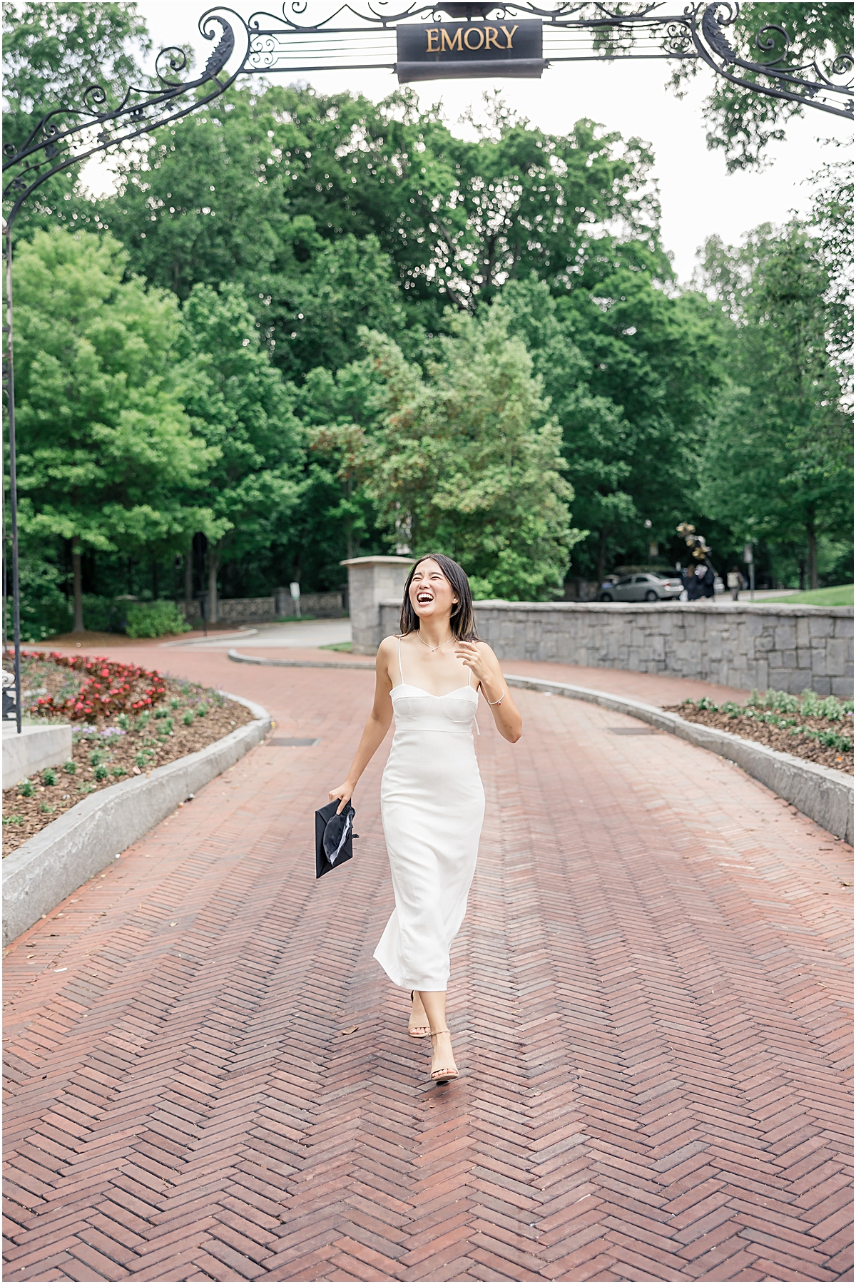 Female college graduate in white dress walking on brick path towards Emory University Senior Photographer holding her black cap for graduation