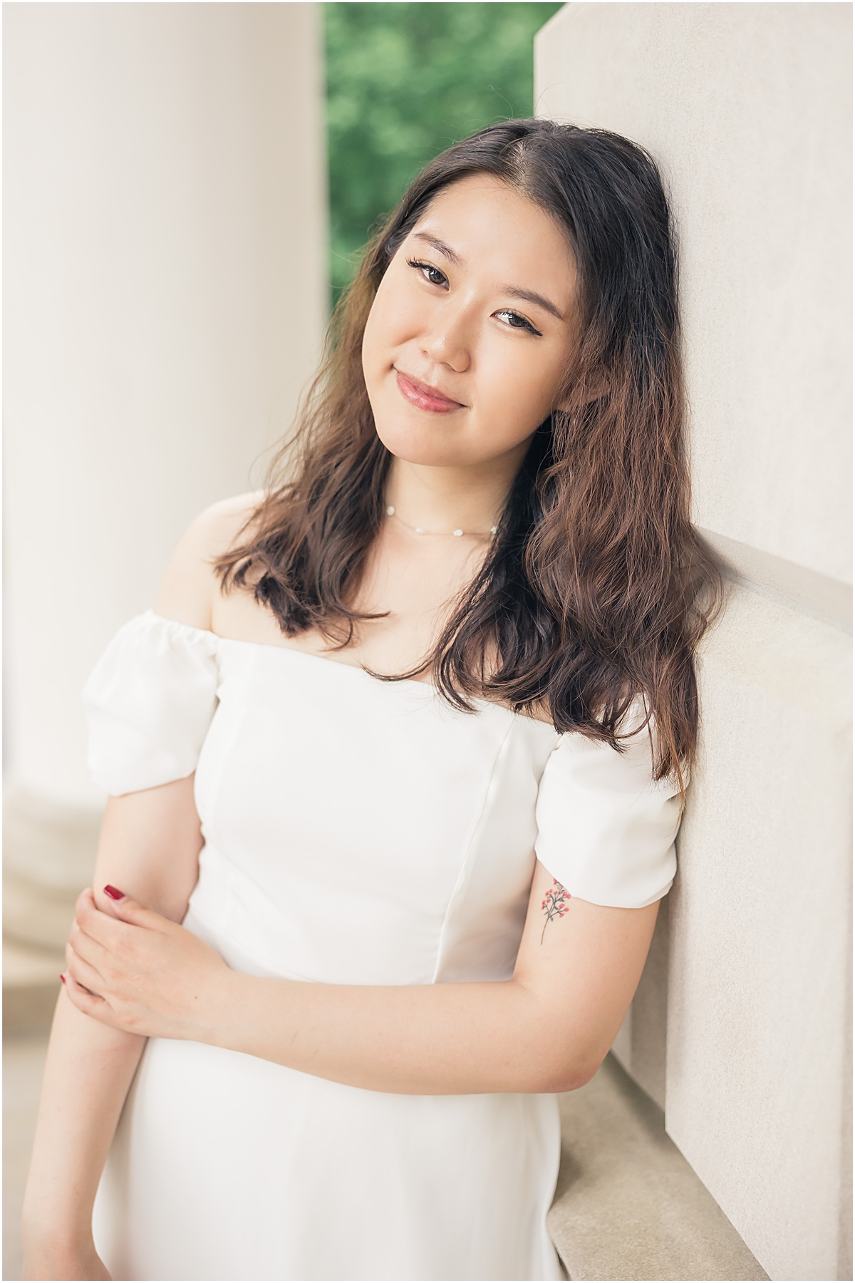 female graduate in white off the shoulder dress leaning against the white wall of a building posing for an Emory University Senior Photographer