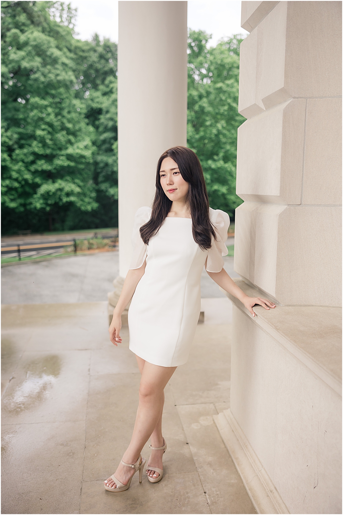 female graduate in white dress leaning against white building looking past Emory University Senior Photographer