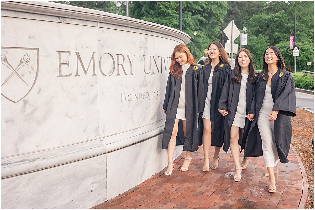 4 female graduates in white dresses and their graduation gowns walking on a brick path in front of an Emory University memorial towards their Emory University Senior Photographer