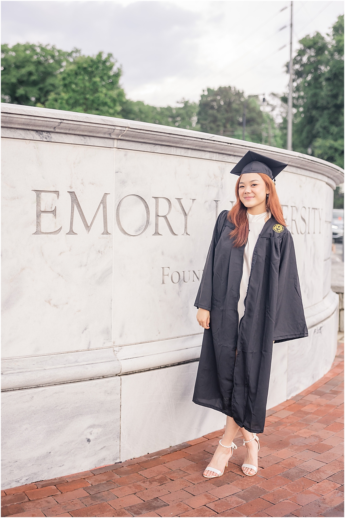 red headed female in a white dress and her black graduation cap & gown smiling at the camera of an Emory University Senior Photographer in front of a stone monument