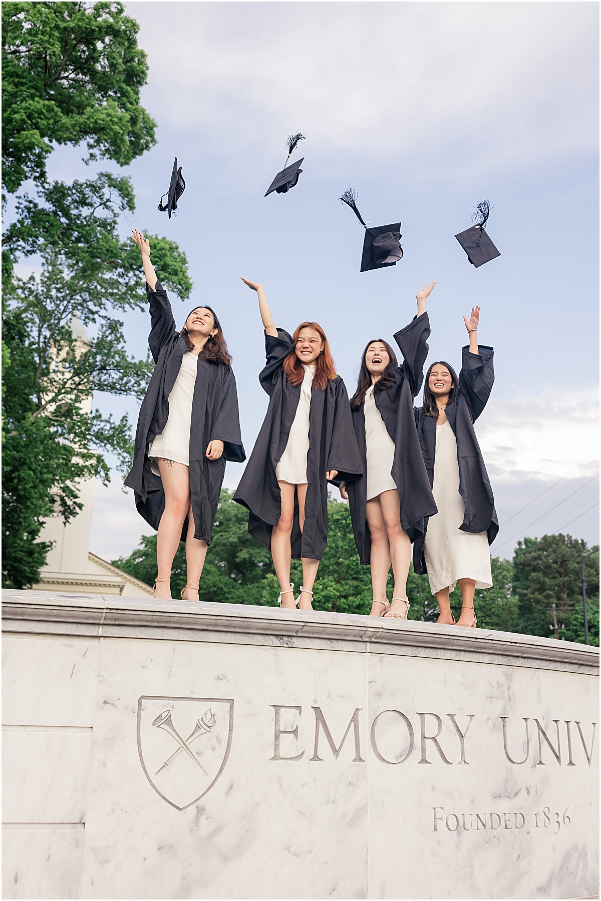 4 female graduates wearing coordinating white dresses and their black graduation gowns and throwing their graduation caps in the air