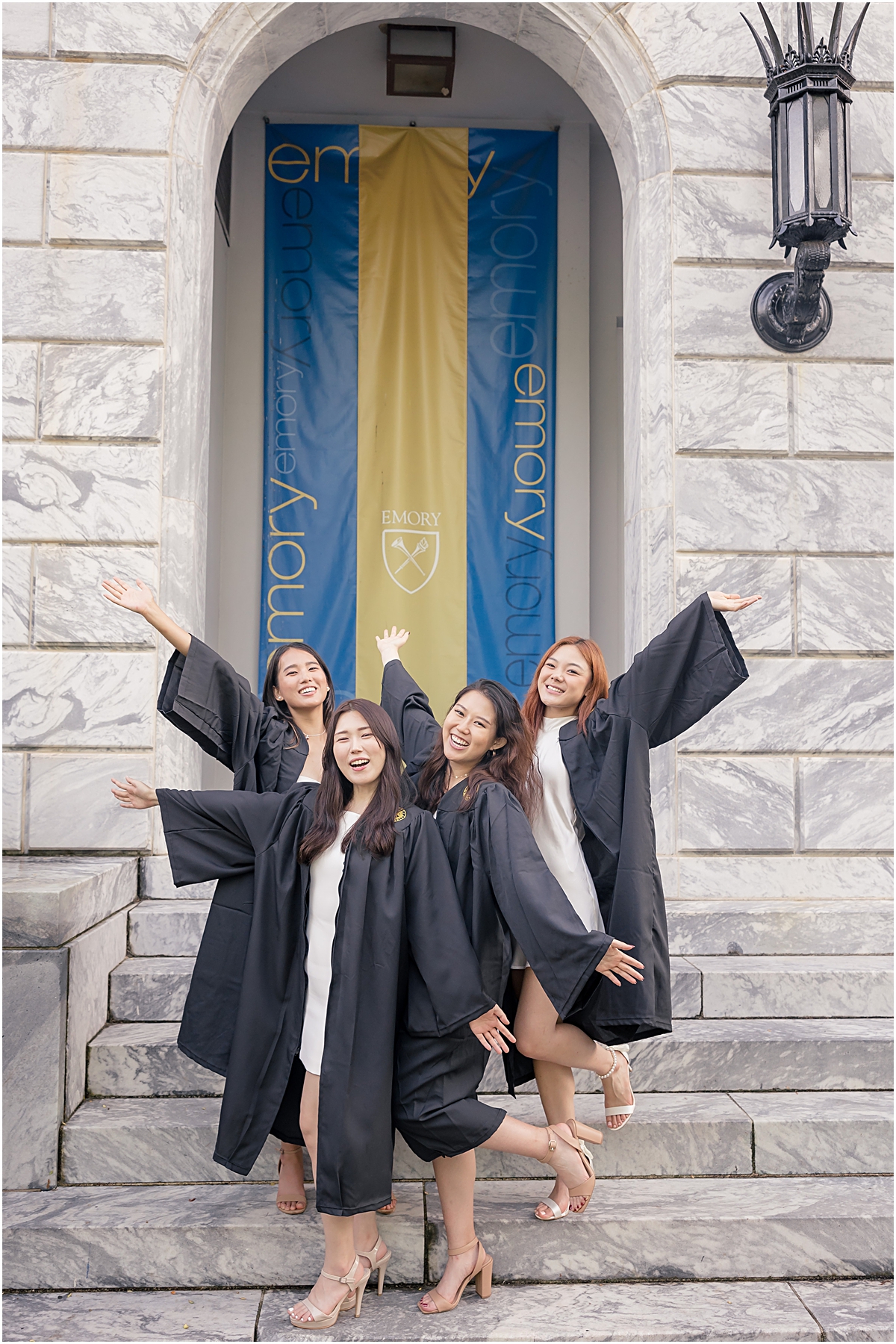 4 Emory University female graduates wearing white dresses and their black graduation gowns posing in front of a blue and gold emory university banner with their hands out stretched