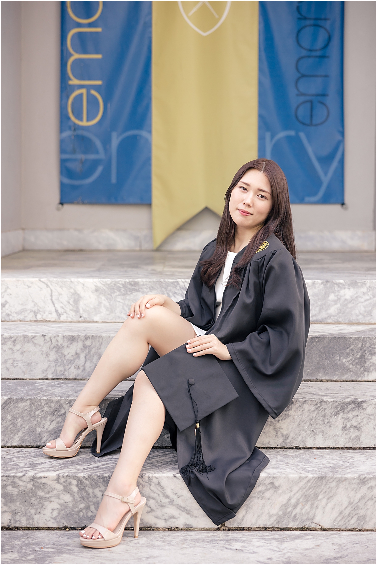 Female sitting on steps in front of gold and blue banner smiling at an Emory University Senior Photographer while she wears her open graduation gown to reveal a white dress and holds her black graduation cap