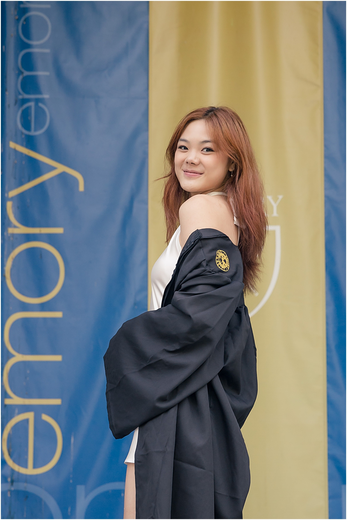 Female standing in front of gold and blue banner smiling at an Emory University Senior Photographer while she wears her open graduation gown off her shoulders to reveal a white dress