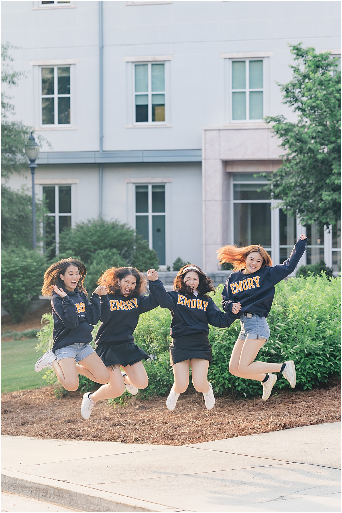 4 female graduates of Emory University in GA jumping in the air wearing matching navy blue long sleeve t-shirts that say "Emory" in block letters in gold with white outlines