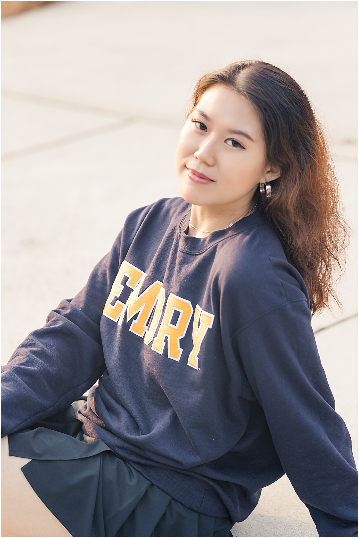Female graduate sitting on the edge of a sidewalk leaned back on her hand wearing a navy blue t-shirt that says "Emory" in yellow block letters with white outlines as she looks up and softly smiles at the Emory University Senior Photographer
