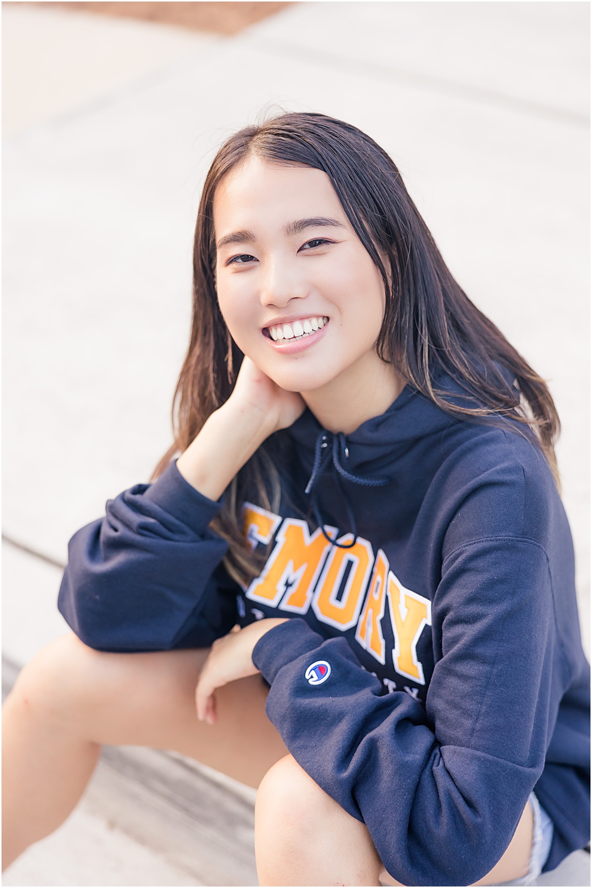 Female graduate sitting on the edge of a sidewalk with her left forearm over her knees and her right elbow propped up on her knee as she leans into her propped hand on the right while wearing a navy blue t-shirt that says "Emory" in yellow block letters with white outlines as she looks up and softly smiles at the Emory University Senior Photographer
