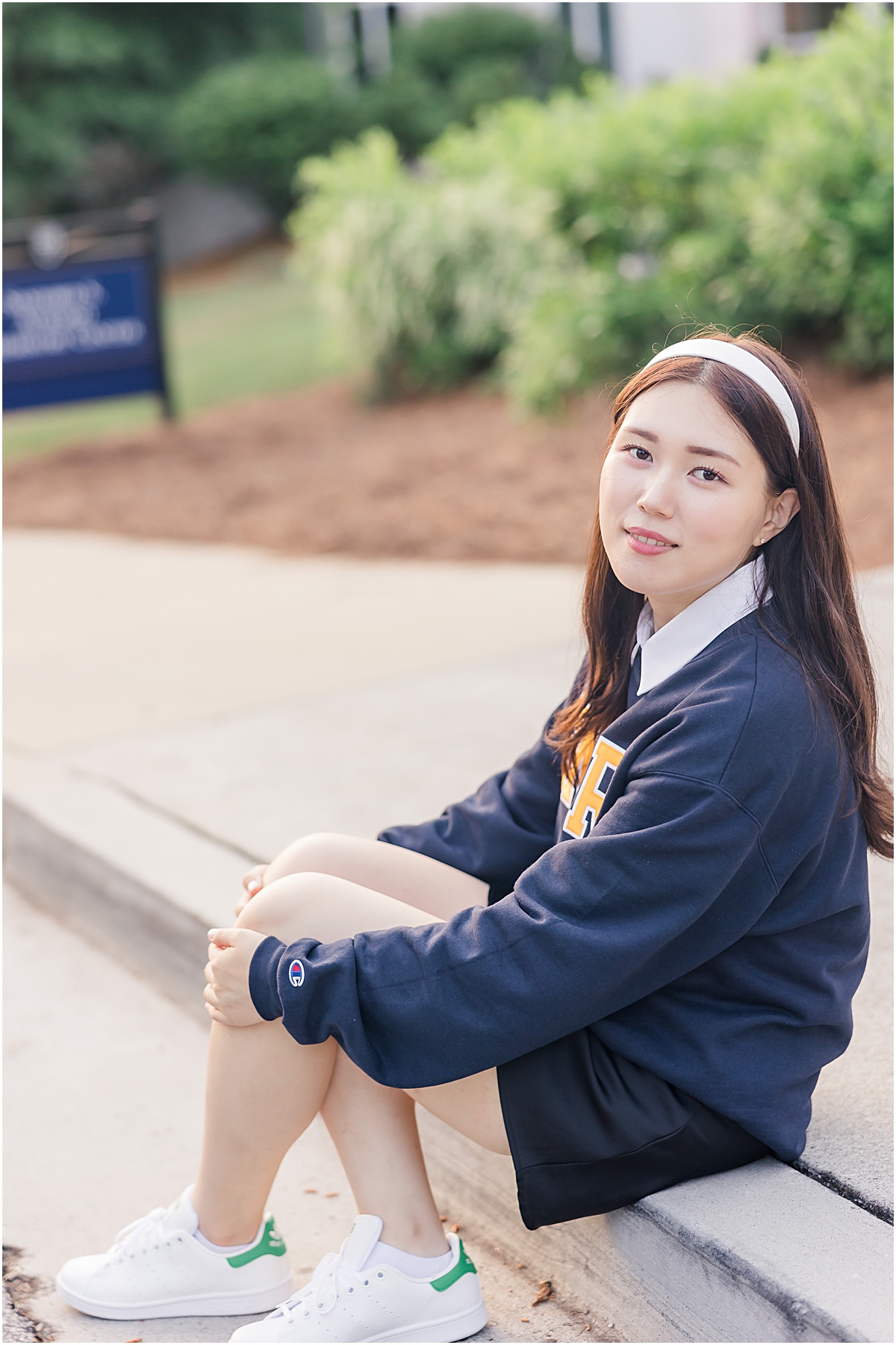 Female graduate sitting on the edge of a sidewalk with her left forearm over her knees and her right elbow propped up on her knee as she holds her bent knees with both hands while wearing a navy blue t-shirt as she looks to her left and softly smiles at the Emory University Senior Photographer